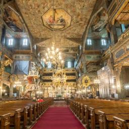 Kodama Foto - Wnętrze zabytkowego, drewnianego kościoła z bogato zdobionym sufitem, ołtarzem i ławkami dla wiernych, oświetlone przez żyrandole i światło dzienne.