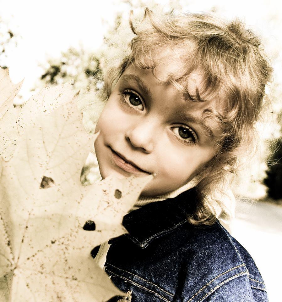 Uśmiechnięta dziewczynka z kręconymi blond włosami, ubrana w dżinsową kurtkę, trzyma przed twarzą duży, jasny liść klonu z dziurami, w sepiowej tonacji.