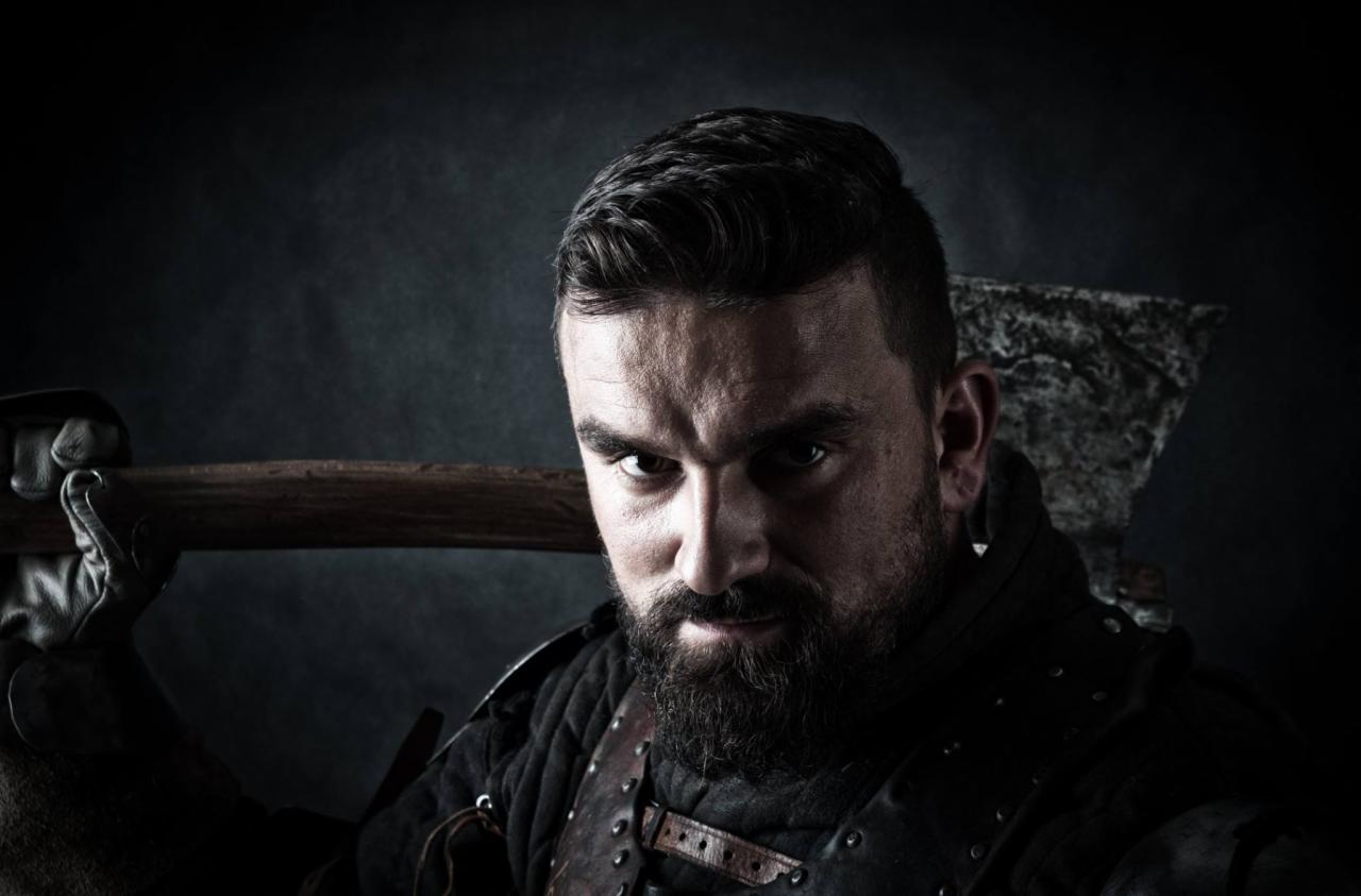 Portret brodatego mężczyzny w skórzanej zbroi, trzymającego topór na ramieniu, z intensywnym spojrzeniem w obiektyw.