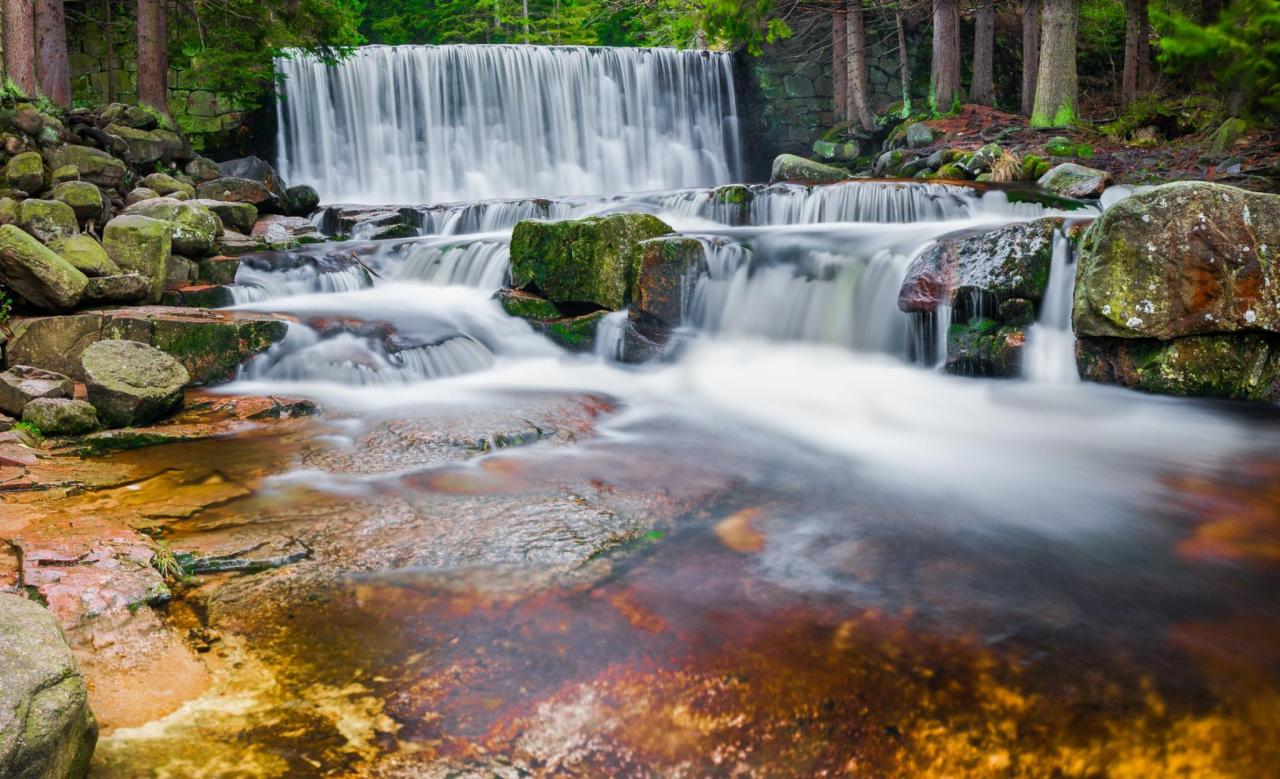 Szeroki wodospad kaskadowy w leśnym otoczeniu, długi czas naświetlania rozmywa wodę, mech porasta kamienie na pierwszym planie.