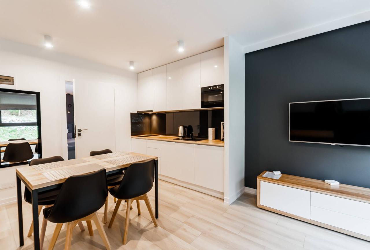 Jasne wnętrze mieszkania z aneksem kuchennym, drewnianym stołem z czarnymi krzesłami, białą zabudową kuchenną z czarnym panelem szklanym i drewnianym blatem, ciemnoszarą ścianą z telewizorem...