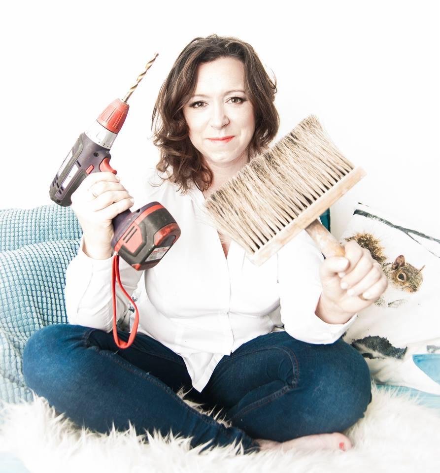 Kobieta siedzi na puszystym dywanie, trzymając w jednej ręce wiertarkę akumulatorową, a w drugiej duży pędzel do tapetowania, uśmiechając się do kamery.