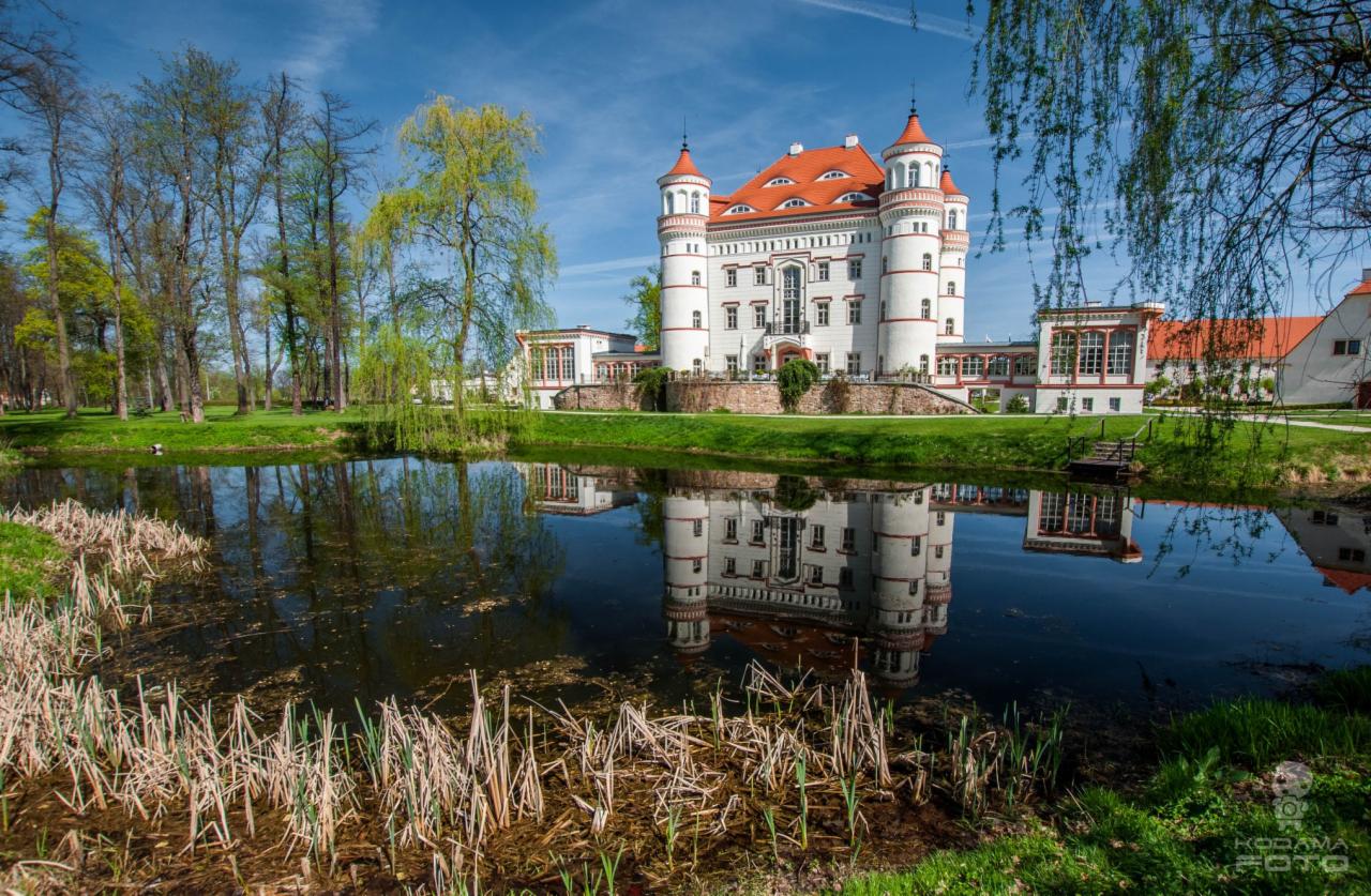 Biały zamek z czerwonym dachem odbijający się w stawie, otoczony zielenią drzew i trawników.