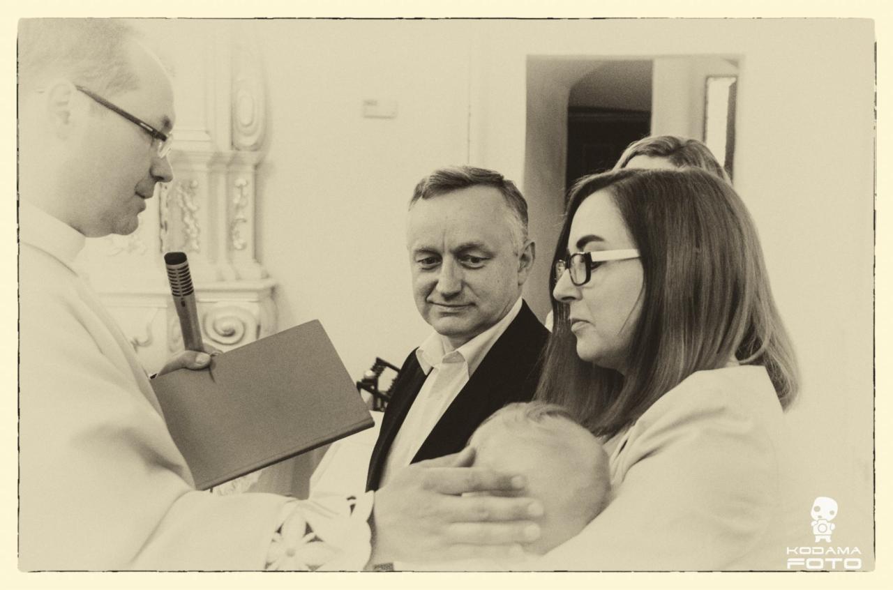 Ksiądz z mikrofonem prowadzi ceremonię, rodzice trzymają dziecko podczas chrztu, stonowana kolorystyka.