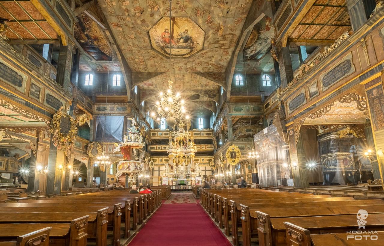 Wnętrze zabytkowego, drewnianego kościoła z bogato zdobionym sufitem, ołtarzem i ławkami dla wiernych, oświetlone przez żyrandole i światło dzienne.