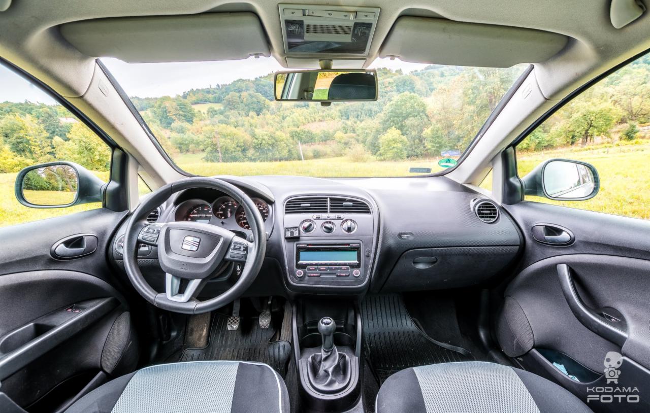 Wnętrze samochodu Seat z widokiem na zielone wzgórza za szybą, deska rozdzielcza z radiem i klimatyzacją, czarna kierownica z logo.