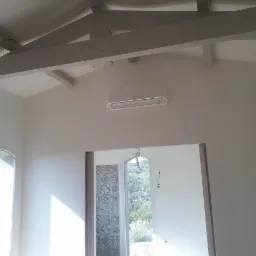 Jasne wnętrze po remoncie z widocznymi drewnianymi belkami stropowymi, klimatyzatorem na ścianie i oknem z widokiem na zieleń.