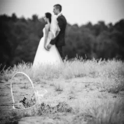 Czarno-białe zdjęcie pary młodej w Łodzi, stojącej na wzgórzu z koszykiem w trawie na pierwszym planie, z rozmytym tłem drzew.