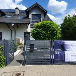 Dom jednorodzinny z panelami fotowoltaicznymi na dachu, klimatyzatorem na elewacji i banerem reklamowym fotowoltaiki i pomp ciepła przy ogrodzeniu.