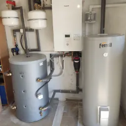 Zestaw urządzeń w kotłowni: biały panel sterowania Panasonic, dwa szare zbiorniki na wodę Galmet i Ferroli, połączone rurami z szarą izolacją.