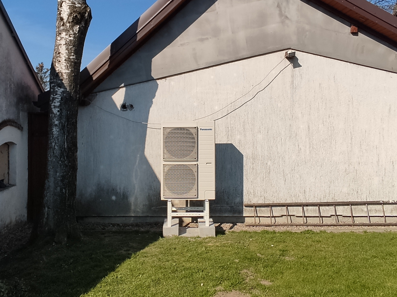 Zewnętrzna jednostka pompy ciepła Panasonic, zamontowana na betonowym fundamencie przy ścianie budynku z widocznym fragmentem drabiny i drzewem brzozy w tle, oświetlona popołudniowym słońcem.