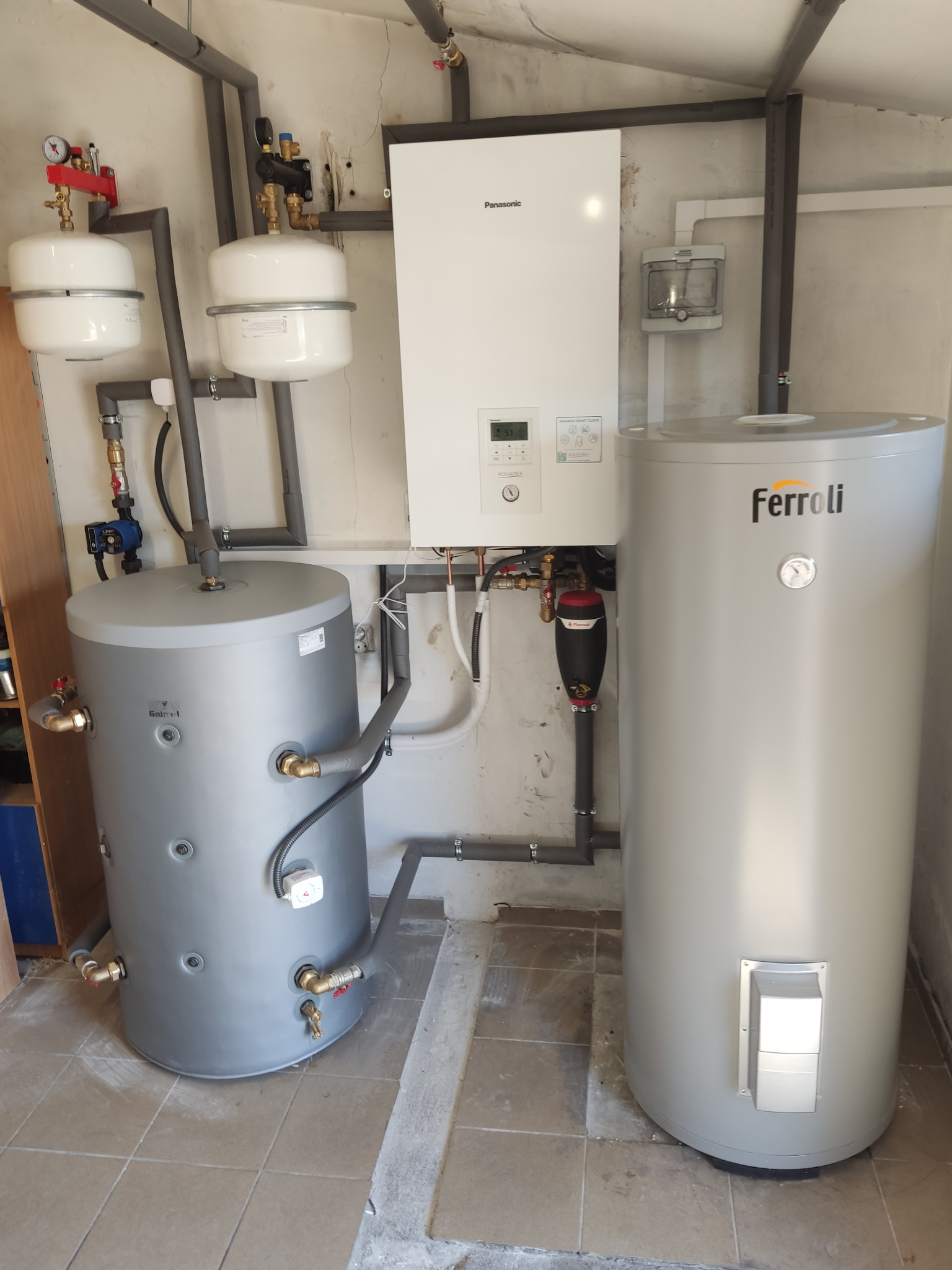 Zestaw urządzeń w kotłowni: biały panel sterowania Panasonic, dwa szare zbiorniki na wodę Galmet i Ferroli, połączone rurami z szarą izolacją.