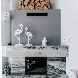 Nowoczesny marmurowy z czarnymi żyłkami z wkładem kominkowym, dekoracjami w postaci białych flamingów i lampionu oraz ułożonym drewnem opałowym na górze.