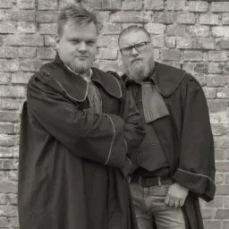 Portret dwóch mężczyzn w togach prawniczych na tle ceglanej ściany, jeden w okularach i jeansach, drugi z założonymi rękoma.