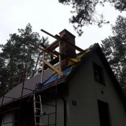 Dom jednorodzinny w trakcie remontu dachu z widocznym kominem, rusztowaniem i elementami drewnianej konstrukcji, zabezpieczony folią, na tle drzew iglastych i pochmurnego nieba.