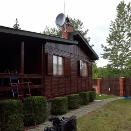 Malowanie drewnianego domu na brązowo z użyciem kompresora; widoczny pracownik na dachu przy kominie i antenie satelitarnej.