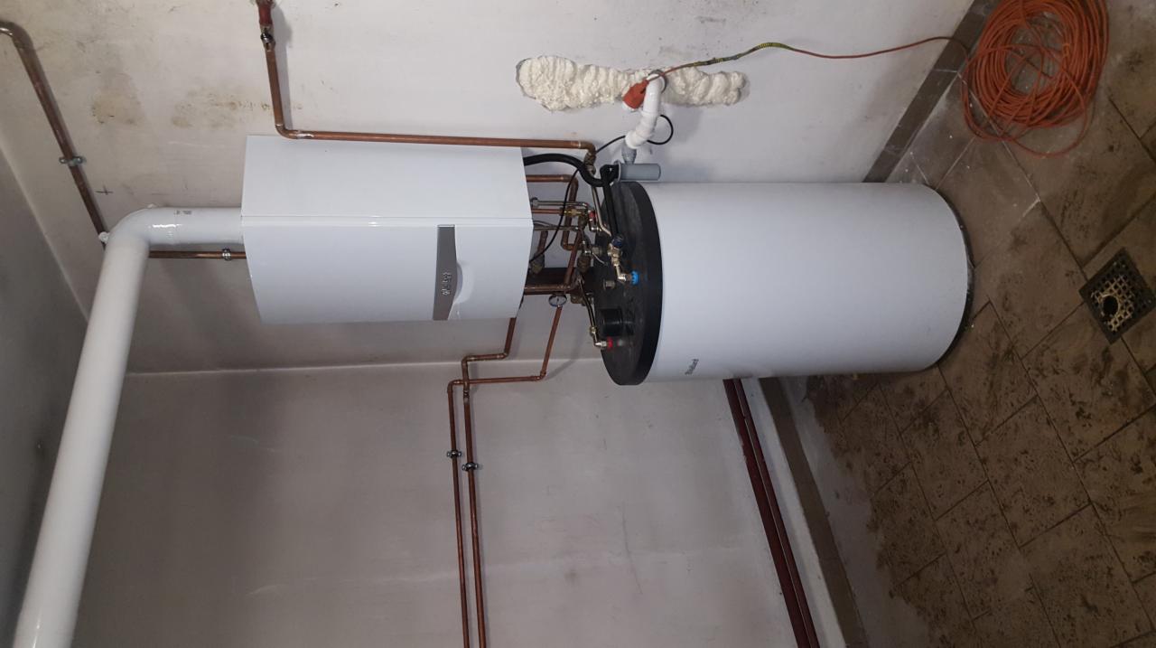 Biały bojler z podłączonymi miedzianymi rurami, widoczne połączenia, izolacja piankowa wokół rur w ścianie i zwój pomarańczowego kabla na podłodze.