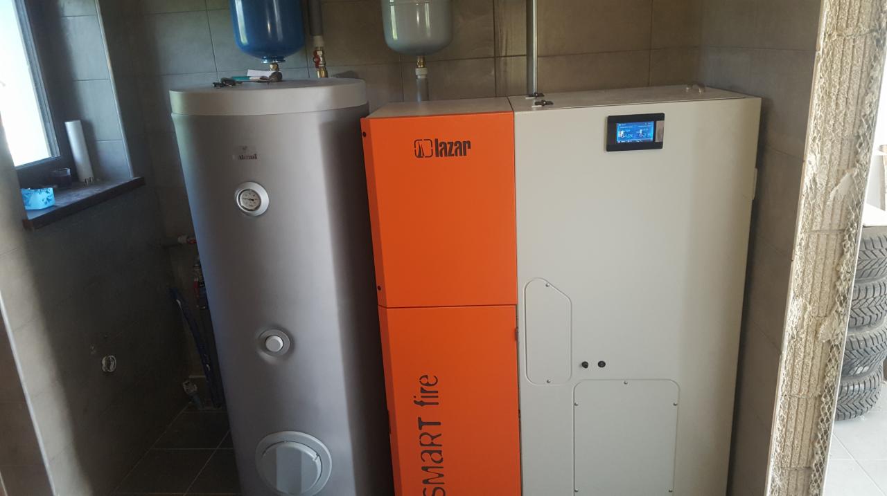 Zestaw urządzeń grzewczych: srebrny zasobnik ciepłej wody i biało-pomarańczowy piec z wyświetlaczem, zainstalowane w pomieszczeniu z szarą glazurą, widoczne opony w tle.