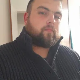 Portret brodatego mężczyzny w szarym swetrze na tle ściany z lustrem.