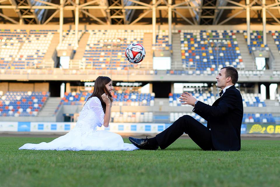 Para młoda siedzi na murawie stadionu, panna młoda w białej sukni ślubnej, pan młody w czarnym smokingu, nad nimi lewituje piłka do nogi.