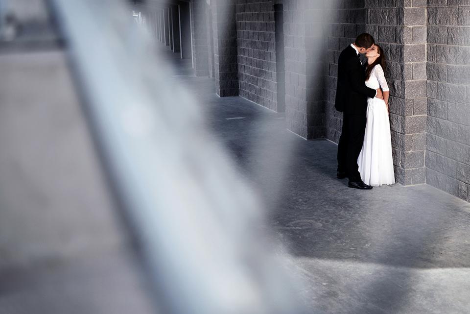 Para narzeczonych w czarno-białej tonacji, pocałunek pod arkadami, on w czarnym garniturze, ona w białej sukni z rękawami 3/4, tło z cegieł.