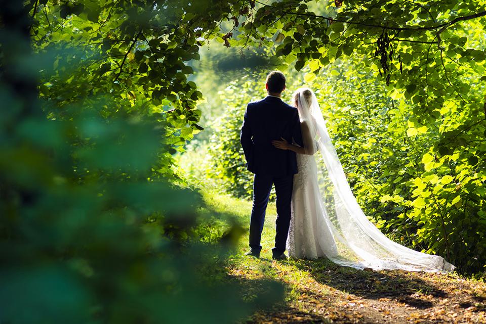 Para młoda, on w granatowym garniturze, ona w białej sukni z długim welonem, stoją tyłem na leśnej ścieżce, otoczeni zielenią drzew i krzewów, w słoneczny dzień.