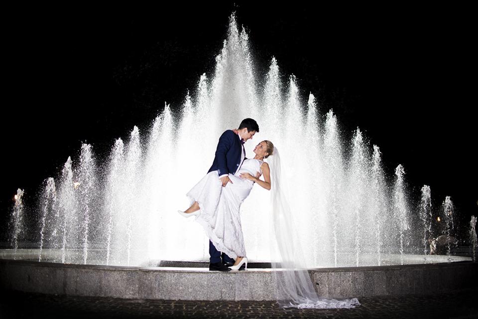 Para młoda w tańcu na tle podświetlonej fontanny w nocy, panna młoda w białej sukni z długim welonem i białych szpilkach, pan młody w granatowym garniturze z muszką.