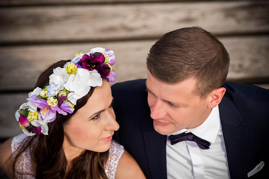 Portret narzeczonych: kobieta w wianku z białych, fioletowych i różowych kwiatów spogląda na mężczyznę w granatowym garniturze i bordowej muszce na tle drewnianych desek.