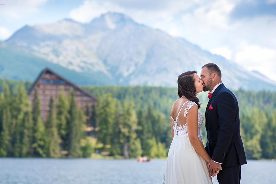Para młoda całuje się na tle górskiego jeziora i zalesionego zbocza z charakterystycznym budynkiem.