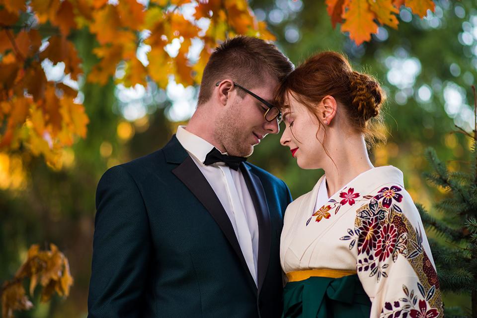 Para młoda w jesiennej scenerii: mężczyzna w granatowym smokingu z muchą, kobieta w zdobionym kimonie, stykają się czołami na tle drzew z pomarańczowymi liśćmi.