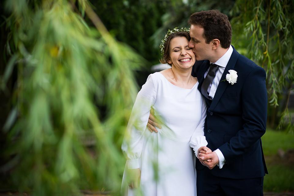 Uśmiechnięta panna młoda w białej sukni z wiankiem na głowie, całowana w policzek przez pana młodego w granatowym garniturze i z białą butonierką, na tle zieleni.