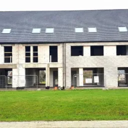 Surowe, szeregowe domy w trakcie budowy, z widocznymi pustakami, otworami okiennymi i dachami z lukarnami, ogrodzone tymczasowym płotem.