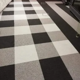 Wzór szachownicy na podłodze z wykładziny dywanowej w odcieniach szarości, z widocznymi włazami rewizyjnymi i fragmentem grzejnika przy ścianie pomalowanej na czerwono.