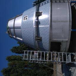 Spaw-mont - Częściowo osłonięty, metalowy zbiornik przemysłowy z widocznym logo producenta, ustawiony na zewnątrz, z drabinką i pracownikiem w tle, na tle błękitnego nieba i drzew.