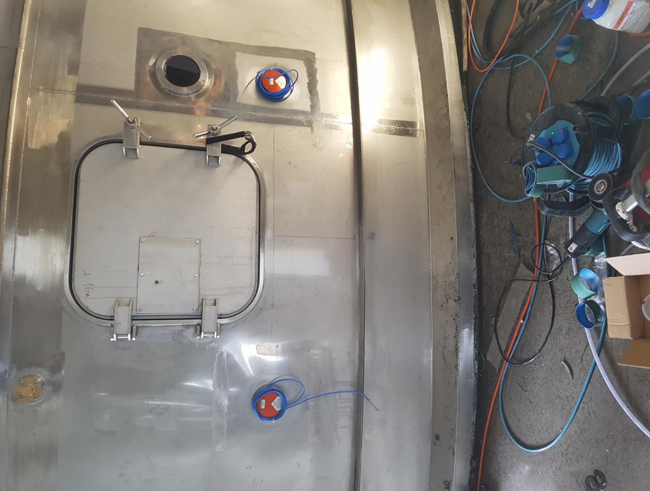 Metalowa konstrukcja zbiornika z włazem inspekcyjnym i elementami instalacji, widoczne ślady spawania i obróbki, narzędzia i kable w tle.
