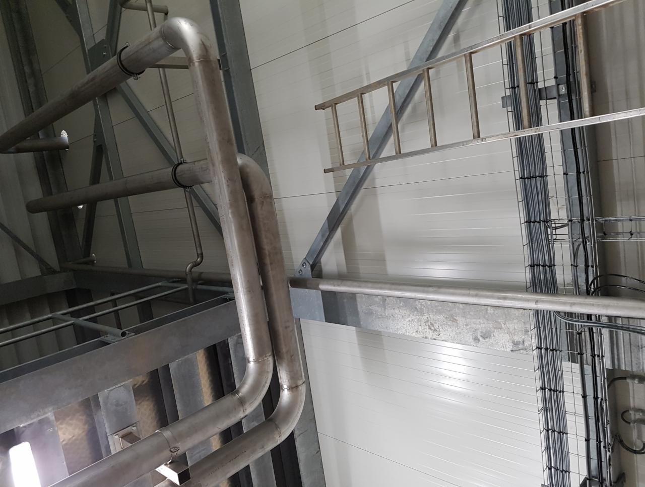 Metalowe rury i konstrukcja wsporcza pod sufitem hali przemysłowej, widoczna drabina i kable w korytkach.