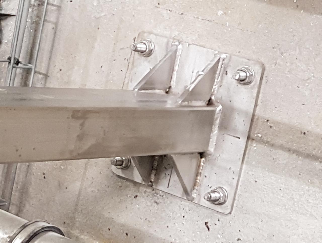 Metalowa konstrukcja wsporcza przyspawana do betonowej powierzchni, widoczne spawy, śruby mocujące i fragment rury w dolnej części kadru.