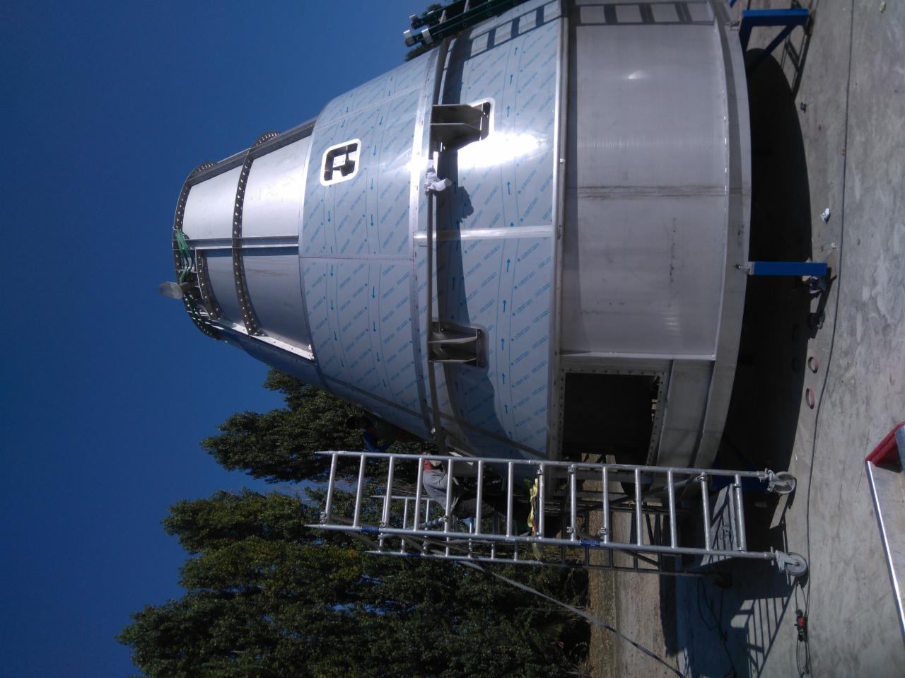 Częściowo osłonięty, metalowy zbiornik przemysłowy z widocznym logo producenta, ustawiony na zewnątrz, z drabinką i pracownikiem w tle, na tle błękitnego nieba i drzew.