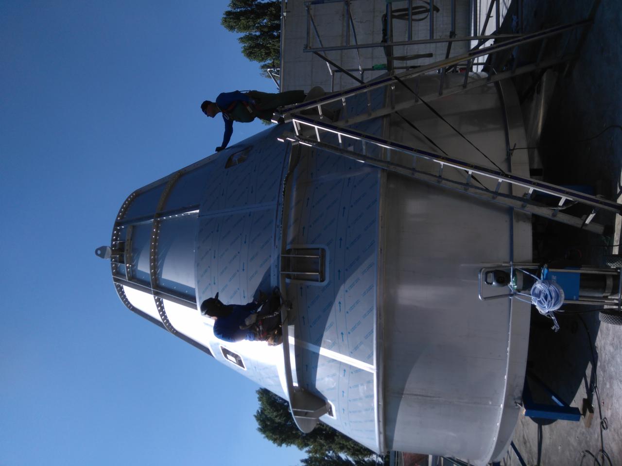 Praca na wysokości: dwóch monterów instaluje metalowe panele na dużej, stożkowej konstrukcji z użyciem rusztowania i narzędzi.