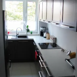 Nowoczesna kuchnia w odcieniach brązu z białym blatem, czarnym zlewem i płytą indukcyjną, oświetlona naturalnym światłem z okna z widokiem na zieleń.