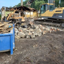 BET GROUNDWORKS SPÓŁKA Z OGRANICZONĄ ODPOWIEDZIALNOŚCIĄ - Żółta koparka Volvo EC220DL podczas rozbiórki budynku, z niebieskim kontenerem na gruz w tle. Widoczne porozrzucane cegły i elementy konstrukcyjne.