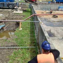 BET GROUNDWORKS SPÓŁKA Z OGRANICZONĄ ODPOWIEDZIALNOŚCIĄ - Instalacja rur na placu budowy z wykopem, widoczny dźwig i pracownik w kasku. Częściowo ogrodzony teren. Prace ziemne w toku, z widocznymi elementami infrastruktury.