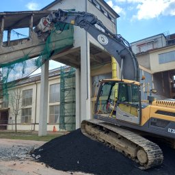 BET GROUNDWORKS SPÓŁKA Z OGRANICZONĄ ODPOWIEDZIALNOŚCIĄ - Żółty koparko-ładowarka z hydraulicznymi nożycami tnie betonowy element konstrukcyjny budynku w trakcie rozbiórki. Widoczne zabezpieczenia siatką.