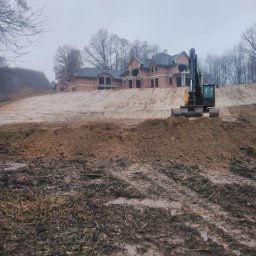 BET GROUNDWORKS SPÓŁKA Z OGRANICZONĄ ODPOWIEDZIALNOŚCIĄ - Żółto-czarna koparka na błotnistym terenie, w tle surowy budynek z czerwonej cegły pod szarym niebem, drzewa bez liści. Etap prac ziemnych.