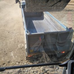 BET GROUNDWORKS SPÓŁKA Z OGRANICZONĄ ODPOWIEDZIALNOŚCIĄ - Kamera z kabiny koparki ukazuje załadunek gruzu do skrzyni wywrotki. W tle widać teren budowy i przesuwające się tumany kurzu.
