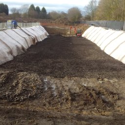 BET GROUNDWORKS SPÓŁKA Z OGRANICZONĄ ODPOWIEDZIALNOŚCIĄ - Wykop zabezpieczony geowłókniną na zboczach, z mini koparką w tle. Prace ziemne w toku, widoczne ślady opon i hałda ziemi. Częściowo pochmurne niebo.