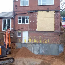 Pomarańczowa koparka Hamilton Plant Hire przy wykopie fundamentów obok ceglanego budynku z zakrytymi otworami okiennymi i żółtymi numerami na ścianie.