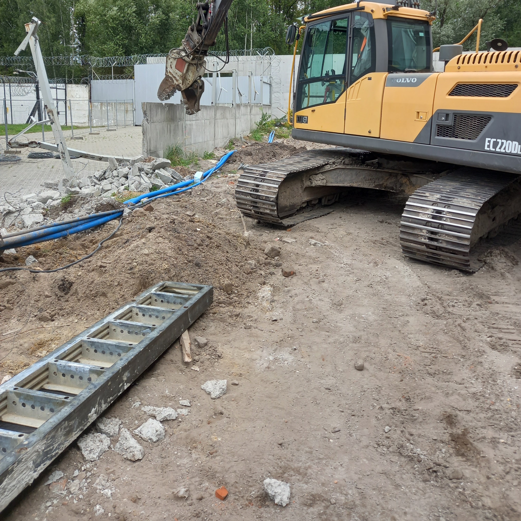 Żółta koparka Volvo EC220DL z mechanicznymi szczękami kruszy betonowy mur. Obok leżą gruz, metalowa drabina i niebieskie rury. W tle ogrodzenie z drutem kolczastym.