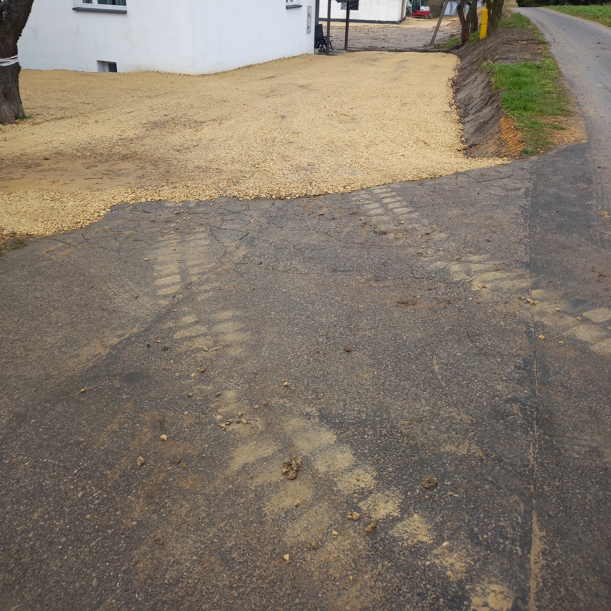 Utwardzony teren żwirem przy białym budynku. Ślady opon na asfalcie, widoczny fragment drogi i trawnika. Na drugim planie koparka.