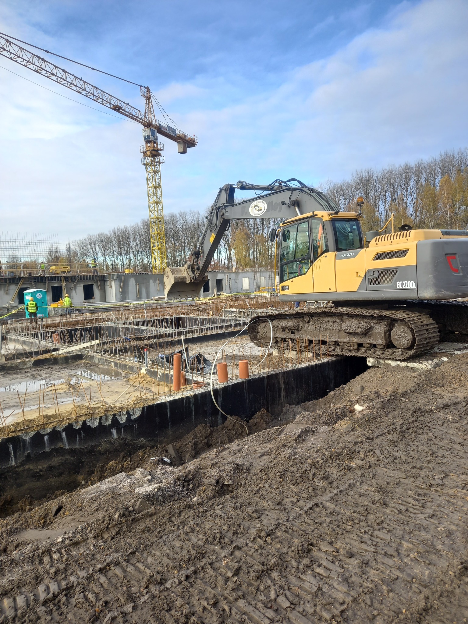 Żółta koparka Volvo EC220DL na placu budowy z widocznym zbrojeniem fundamentów, rurami drenażowymi i żurawiem w tle, w pochmurny dzień.